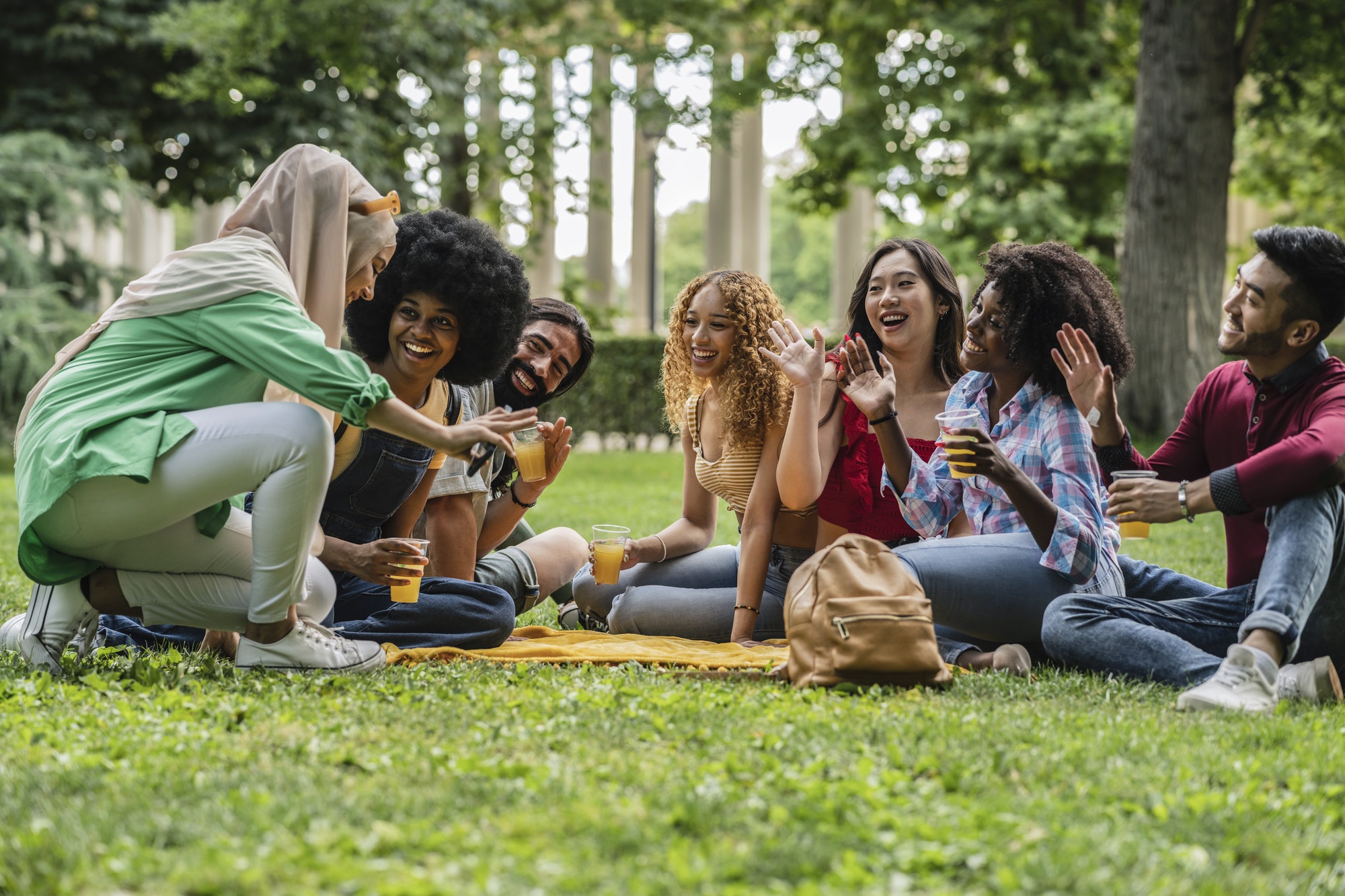 Multiracial group of friends sitting on the grass in a public park drinking orange juice and waving